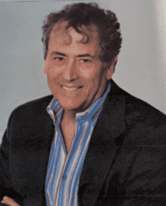 A smiling man with curly hair in a dark blazer and striped shirt.